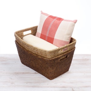 May include: Two rectangular woven baskets, one inside the other, with handles. The top basket contains a rolled cream blanket and a red and white plaid cushion. The baskets are on a white wooden surface.