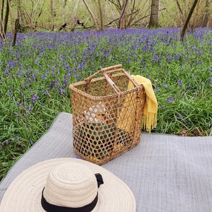 K&ouml;nnte beinhalten: Eine Picknick-Szene mit einem Weidenkorb, einem breitkrempigen Strohhut mit schwarzem Band und einem gelben Schal. Der Korb steht auf einer grauen Decke, mit einem Hintergrund aus lila Blumen und B&auml;umen.