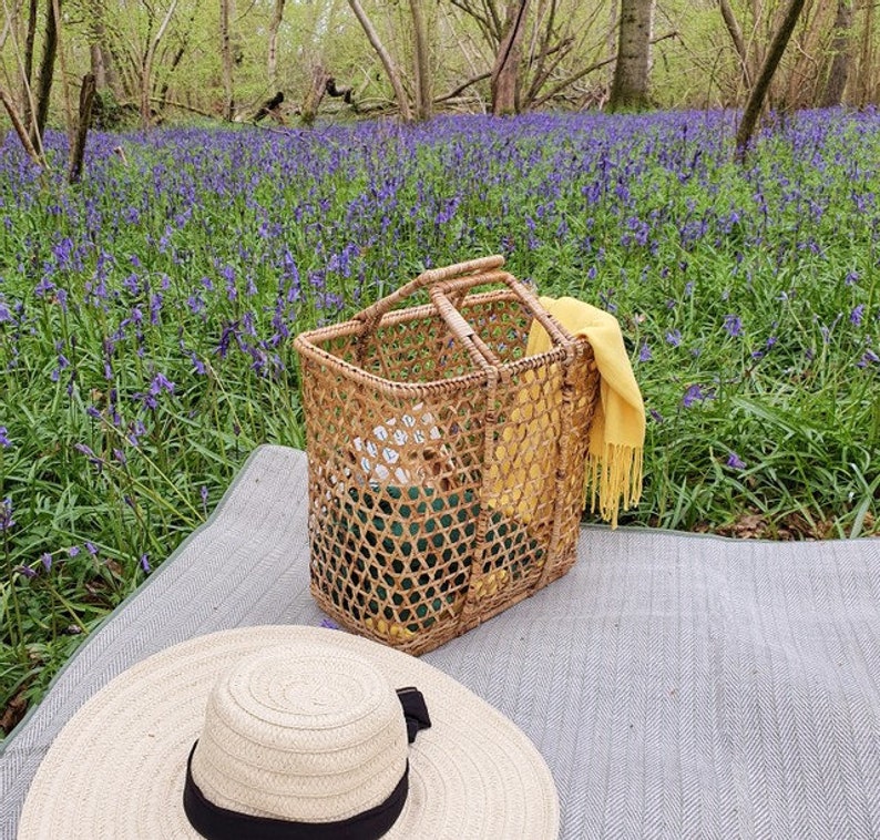 K&ouml;nnte beinhalten: Eine Picknick-Szene mit einem Weidenkorb, einem breitkrempigen Strohhut mit schwarzem Band und einem gelben Schal. Der Korb steht auf einer grauen Decke, mit einem Hintergrund aus lila Blumen und B&auml;umen.