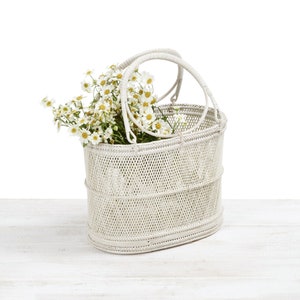 Pu&ograve; includere: Un cesto bianco intrecciato con due manici, pieno di fiori bianchi e gialli. Il cesto ha una forma ovale e un motivo strutturato. I fiori sono margherite e il cesto &egrave; su una superficie di legno bianco.