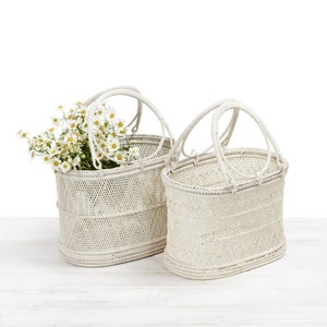 Pu&ograve; includere: Due cesti in vimini bianco sporco con manici ad arco. Un cesto contiene un bouquet di fiori bianchi e gialli. I cesti sono di forma ovale e poggiano su una superficie di legno bianco.
