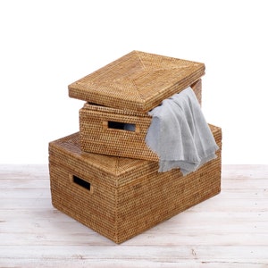 May include: Three woven wicker storage boxes stacked on top of each other. The top box is open and a grey fabric is inside. The boxes are on a white wooden surface.