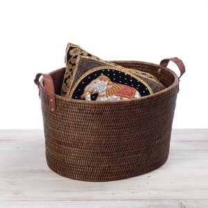 May include: A brown wicker basket with leather handles, containing a black and gold embroidered cushion with an elephant design.