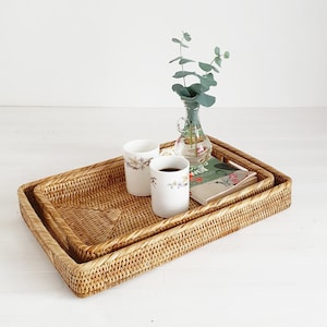 May include: Two white ceramic mugs with floral designs sit on a woven rattan tray with a book and a glass vase with green eucalyptus stems.