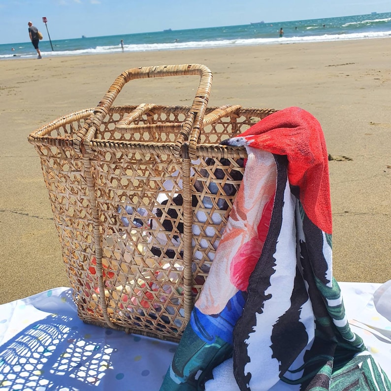 K&ouml;nnte beinhalten: Ein geflochtener Weidenkorb mit Henkel steht auf einer Stranddecke. Ein farbenfrohes Strandtuch mit roten, wei&szlig;en und gr&uuml;nen Blumenmustern h&auml;ngt &uuml;ber der Seite des Korbes. Der Korb ist mit Gegenst&auml;nden gef&uuml;llt, was auf einen Strandtag hindeutet.