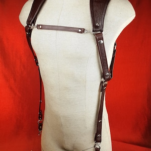 May include: A dark brown leather camera harness featuring wide shoulder straps, a horizontal chest strap, and silver-toned metal hardware. The harness includes adjustable vertical straps with swivel clips for attaching a camera, displayed on a mannequin.