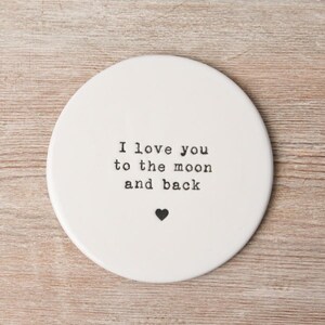 May include: White ceramic coaster with the text "I love you to the moon and back" in black typewriter font. A small black heart is below the text.
