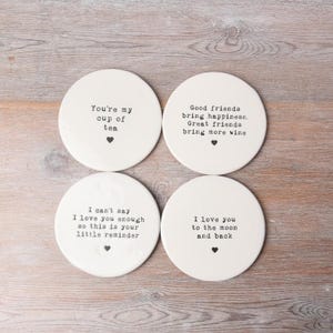 May include: Set of four white ceramic coasters with black text. The coasters feature different sayings, including "You're my cup of tea", "Good friends bring happiness. Great friends bring more wine", "I can't say I love you enough so this is your little reminder", and "I love you to the moon and back".
