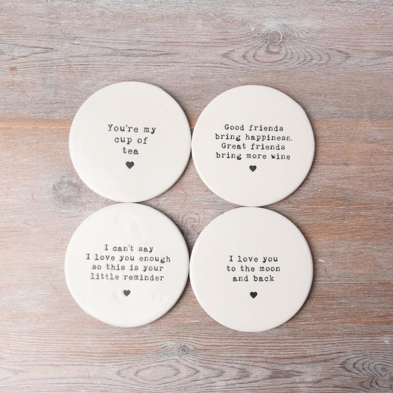 May include: Set of four white ceramic coasters with black text. The coasters feature different sayings, including "You're my cup of tea", "Good friends bring happiness. Great friends bring more wine", "I can't say I love you enough so this is your little reminder", and "I love you to the moon and back".