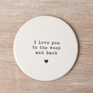 May include: White ceramic coaster with the text "I love you to the moon and back" in black typewriter font. A small black heart is below the text.