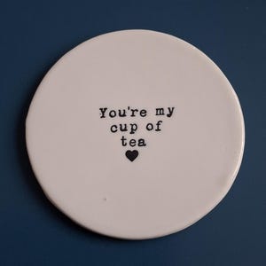 May include: White ceramic coaster with the text "You're my cup of tea" and a small black heart.