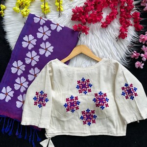 Puede incluir: Blusa color crema de manga larga con dise&ntilde;os florales bordados en rojo y azul. Se ve una bufanda morada con estampados florales blancos. La blusa est&aacute; colgada en una percha de madera, mostrando los detalles.