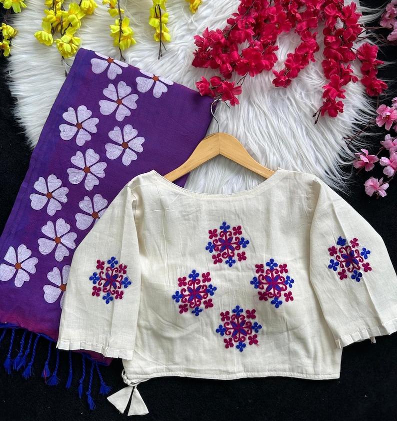 Puede incluir: Blusa color crema de manga larga con dise&ntilde;os florales bordados en rojo y azul. Se ve una bufanda morada con estampados florales blancos. La blusa est&aacute; colgada en una percha de madera, mostrando los detalles.