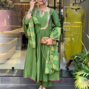 May include: A woman wearing a green ensemble, including a long-sleeved dress with embroidery, matching pants, and a scarf. The dress has pink trim and is paired with gold sandals and a small, patterned clutch. A similar outfit is displayed in the background.