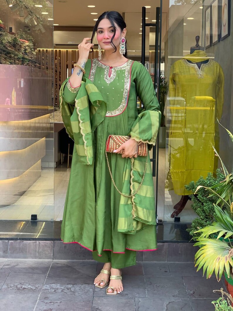May include: A woman wearing a green ensemble, including a long-sleeved dress with embroidery, matching pants, and a scarf. The dress has pink trim and is paired with gold sandals and a small, patterned clutch. A similar outfit is displayed in the background.