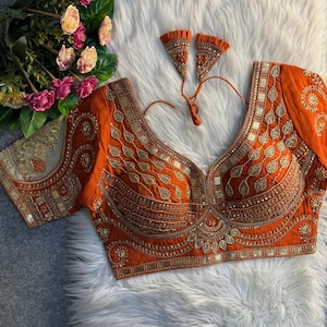 May include: An orange blouse with detailed gold embroidery and embellishments. The blouse has a deep V-neck and short sleeves with intricate patterns. Two matching orange and gold decorative pieces are included.