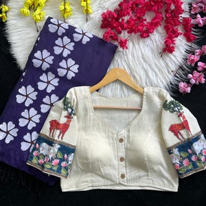 Puede incluir: Blusa color crema con cuello cuadrado y tres botones de madera. Las mangas presentan un dise&ntilde;o de ciervo y &aacute;rbol, con un borde azul y floral. Tambi&eacute;n se ve una tela azul oscuro con estampados florales blancos.