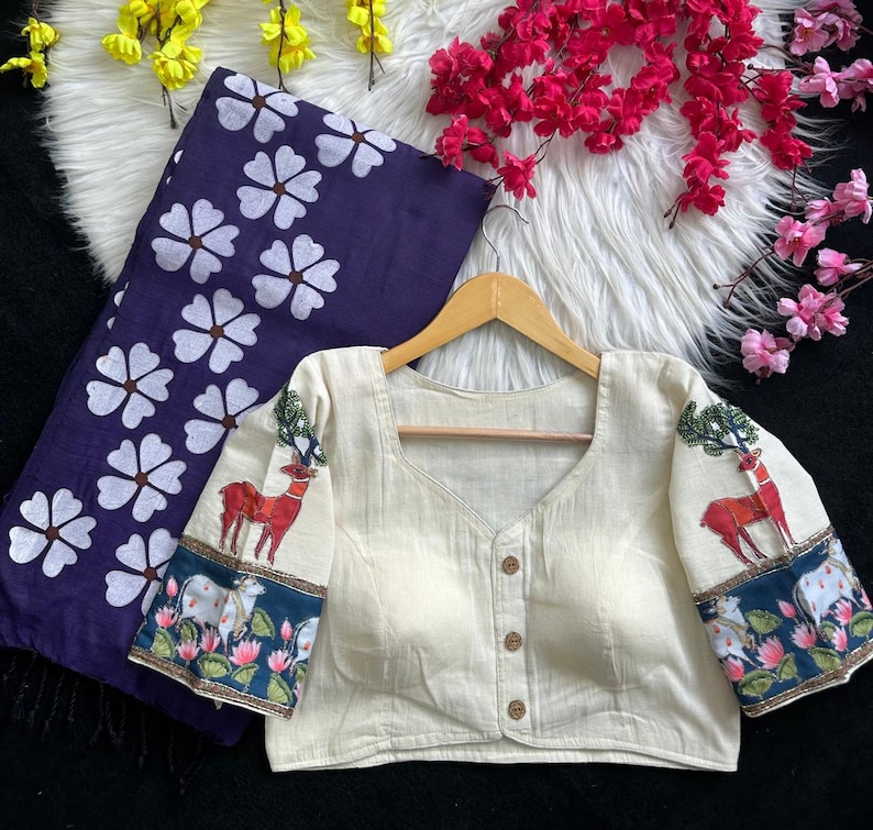 Puede incluir: Blusa color crema con cuello cuadrado y tres botones de madera. Las mangas presentan un dise&ntilde;o de ciervo y &aacute;rbol, con un borde azul y floral. Tambi&eacute;n se ve una tela azul oscuro con estampados florales blancos.