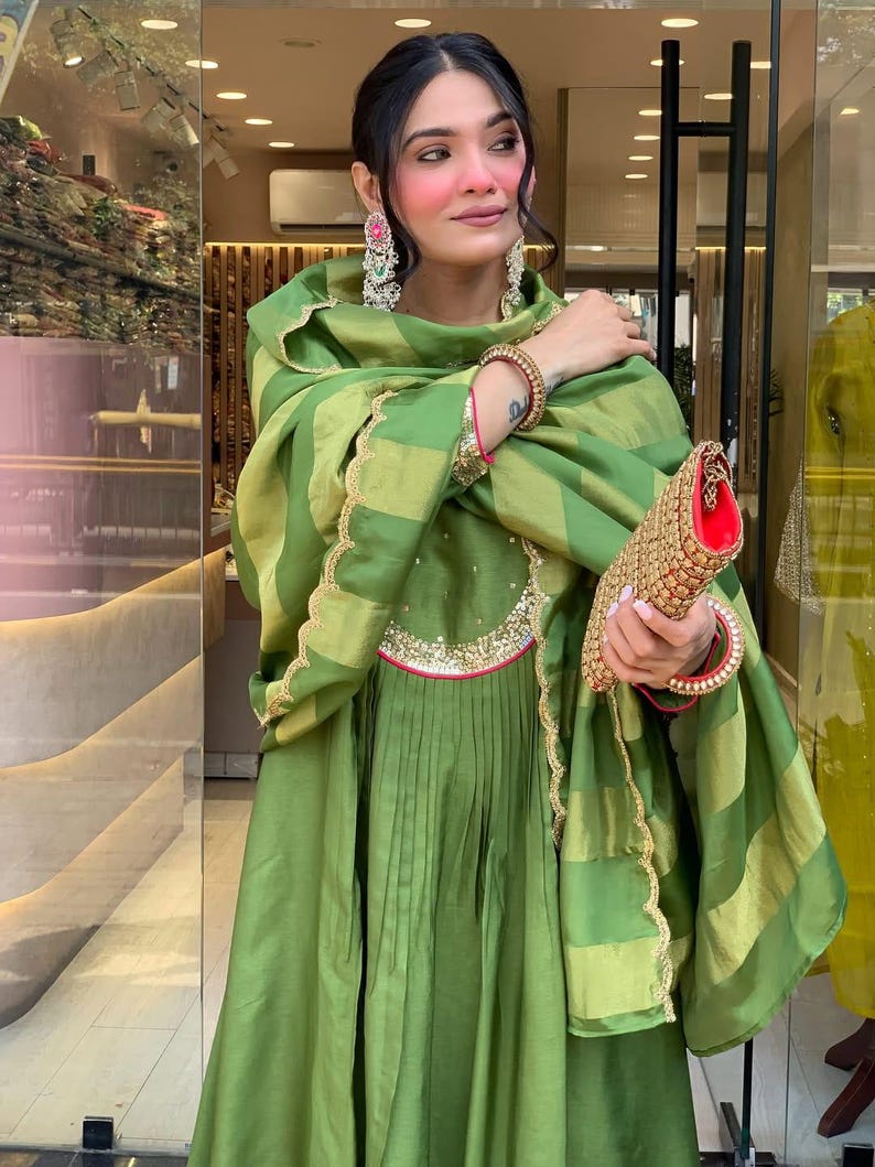 May include: A woman wearing a green dress and shawl with gold trim. The dress has pleats and a decorative neckline. She is holding a gold clutch with a red interior and wearing large earrings and bracelets.