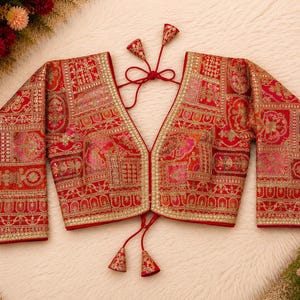 May include: A red long-sleeve blouse featuring an intricate gold and multi-colored patchwork embroidery pattern. The garment has a deep V-neckline, a front opening, and thin red tie closures at the neck and waist, each finished with decorative tassels.