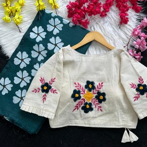 Puede incluir: Blusa crema de manga larga con bordados florales en negro, amarillo y rosa. La blusa se muestra en una percha de madera, junto a una tela verde oscuro con estampados florales blancos. La imagen sugiere ropa hecha a mano.