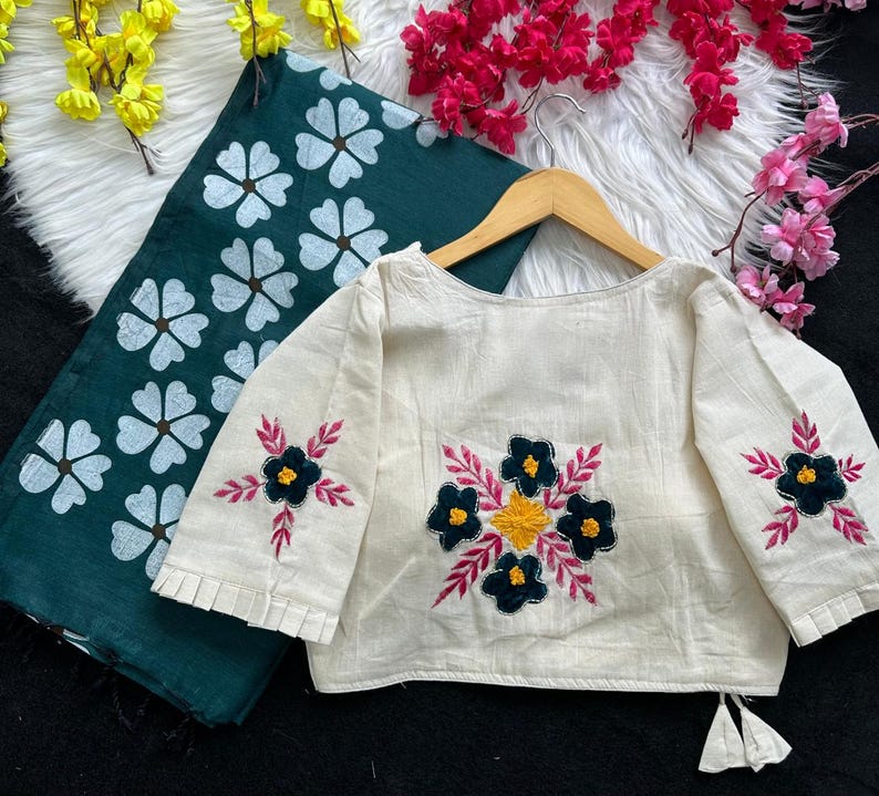 Puede incluir: Blusa crema de manga larga con bordados florales en negro, amarillo y rosa. La blusa se muestra en una percha de madera, junto a una tela verde oscuro con estampados florales blancos. La imagen sugiere ropa hecha a mano.