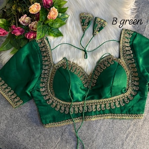 May include: A green blouse with gold embroidery around the neckline, sleeves, and bodice. The blouse features a square neckline and short sleeves. The text "B green" is visible. The blouse is displayed on a white, fluffy surface, with floral accents.