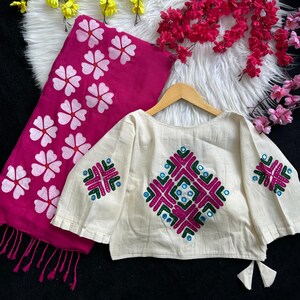 Puede incluir: Una blusa color crema con patrones geom&eacute;tricos bordados en rosa, verde y azul. Una bufanda rosa a juego con dise&ntilde;os florales blancos est&aacute; al lado de la blusa. La blusa est&aacute; en una percha de madera.