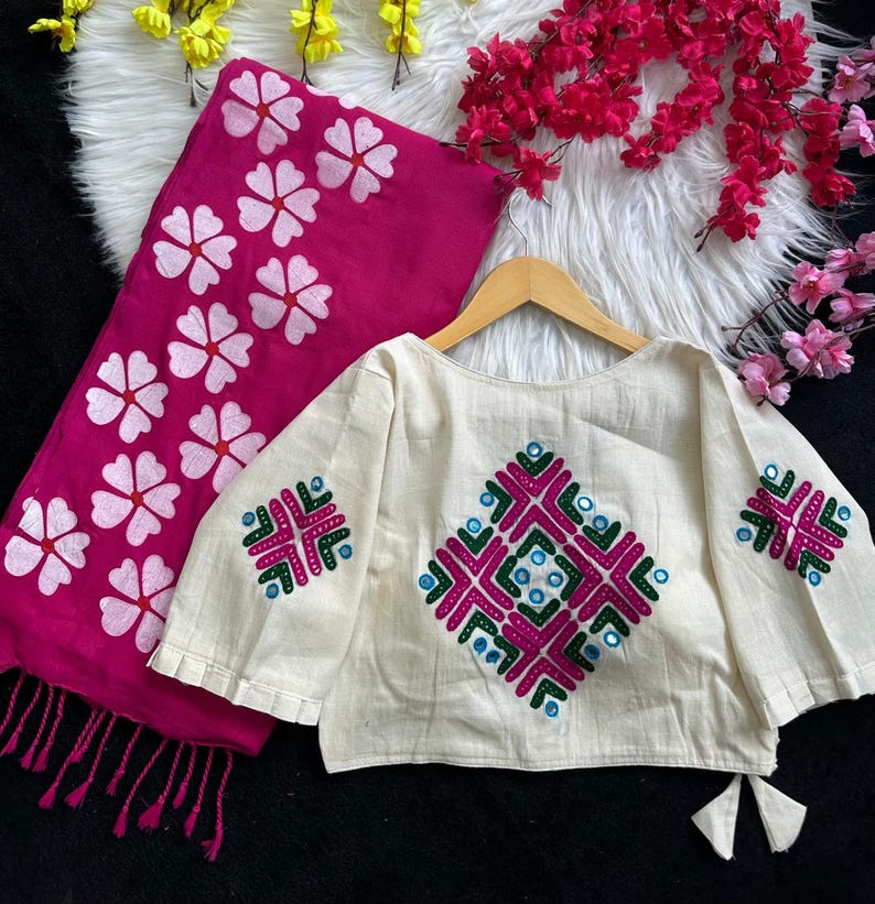 Puede incluir: Una blusa color crema con patrones geom&eacute;tricos bordados en rosa, verde y azul. Una bufanda rosa a juego con dise&ntilde;os florales blancos est&aacute; al lado de la blusa. La blusa est&aacute; en una percha de madera.