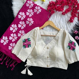Puede incluir: Blusa color crema con detalles bordados en las mangas, combinada con una bufanda fucsia con estampados florales blancos. La blusa tiene cuello en V y cierre de botones. La bufanda tiene flecos.