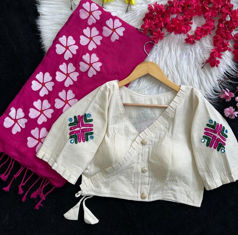 Puede incluir: Blusa color crema con detalles bordados en las mangas, combinada con una bufanda fucsia con estampados florales blancos. La blusa tiene cuello en V y cierre de botones. La bufanda tiene flecos.