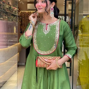 May include: An olive green dress with gold and pink trim, featuring a decorative neckline and long sleeves. The dress is paired with a red and gold clutch. The outfit is accessorized with gold bracelets and large, colorful earrings. The dress has a pleated skirt.