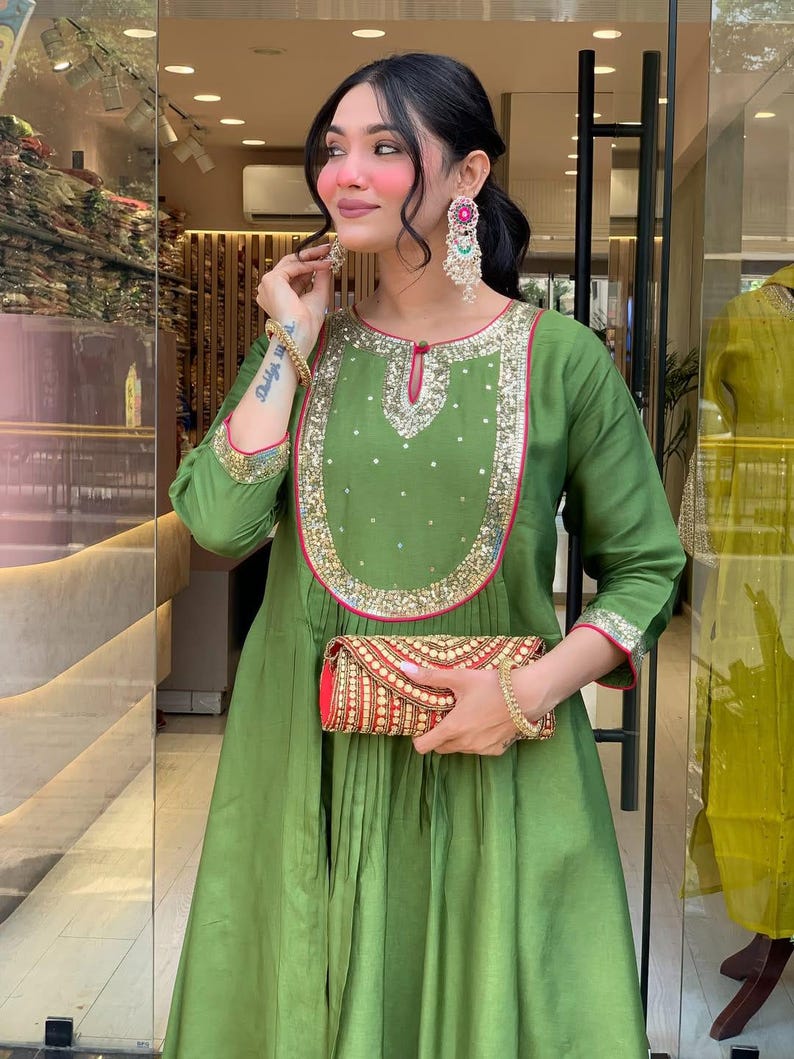 May include: An olive green dress with gold and pink trim, featuring a decorative neckline and long sleeves. The dress is paired with a red and gold clutch. The outfit is accessorized with gold bracelets and large, colorful earrings. The dress has a pleated skirt.