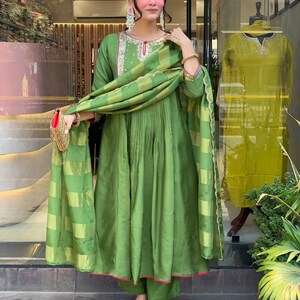 May include: A woman wearing a green silk salwar kameez with a matching dupatta. The outfit features a round neckline with embroidery and pink trim. The dupatta has a striped pattern. She is wearing gold sandals.