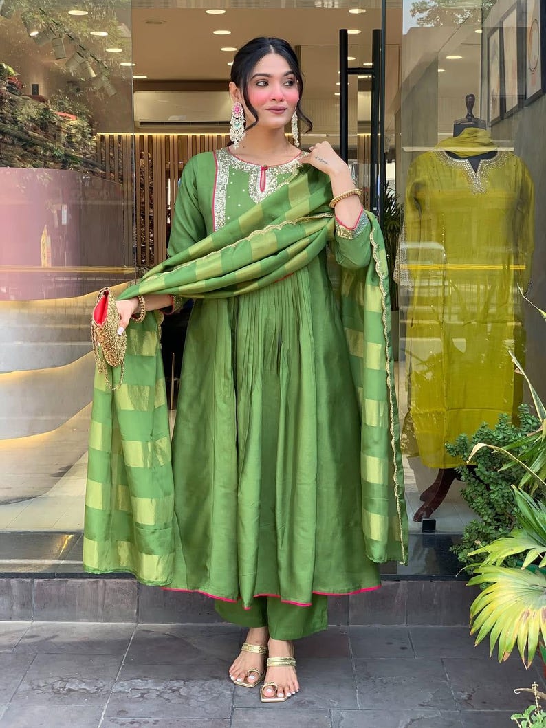 May include: A woman wearing a green silk salwar kameez with a matching dupatta. The outfit features a round neckline with embroidery and pink trim. The dupatta has a striped pattern. She is wearing gold sandals.
