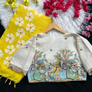 Puede incluir: Una blusa color crema con mangas hasta el codo y una escena bordada detallada de figuras y &aacute;rboles. Una bufanda amarilla con estampados florales blancos est&aacute; al lado de la blusa. La blusa tiene un ribete de encaje festoneado.