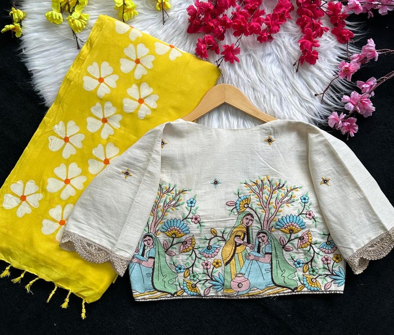Puede incluir: Una blusa color crema con mangas hasta el codo y una escena bordada detallada de figuras y &aacute;rboles. Una bufanda amarilla con estampados florales blancos est&aacute; al lado de la blusa. La blusa tiene un ribete de encaje festoneado.