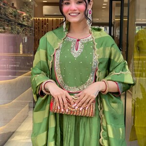 May include: A woman wearing a green ensemble with gold and red accents. The outfit includes a long-sleeved top with intricate embroidery, a matching scarf, and a flowing skirt. She holds a decorative clutch. The ensemble is suitable for special occasions.
