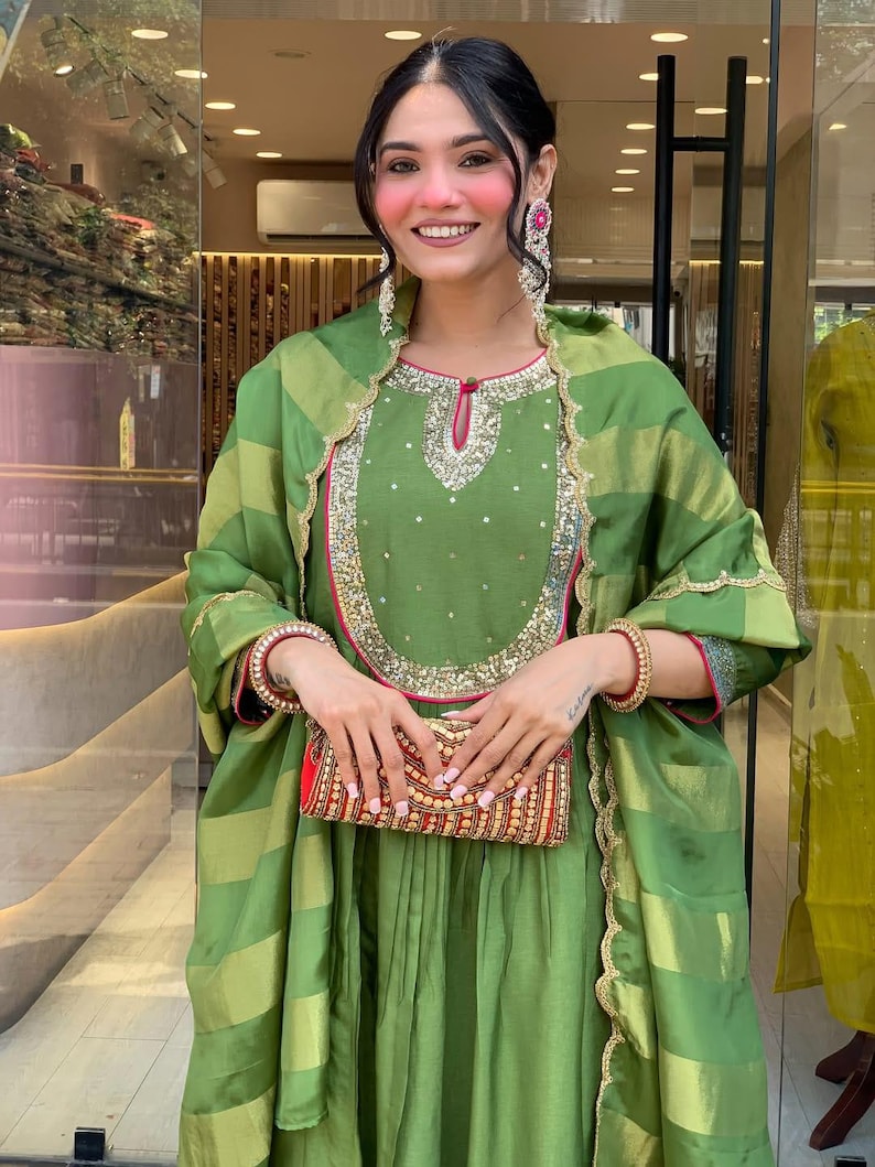 May include: A woman wearing a green ensemble with gold and red accents. The outfit includes a long-sleeved top with intricate embroidery, a matching scarf, and a flowing skirt. She holds a decorative clutch. The ensemble is suitable for special occasions.