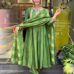May include: A green ensemble featuring a long-sleeved tunic with intricate embroidery around the neckline, paired with matching pants and a draped scarf. The outfit has pink trim and is accessorized with gold sandals and a small purse.