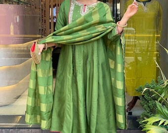 May include: A green ensemble featuring a long-sleeved tunic with intricate embroidery around the neckline, paired with matching pants and a draped scarf. The outfit has pink trim and is accessorized with gold sandals and a small purse.