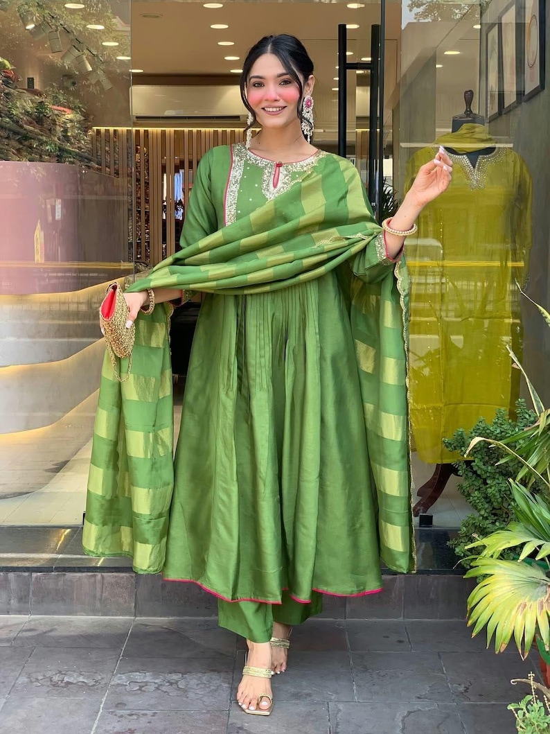 May include: A green ensemble featuring a long-sleeved tunic with intricate embroidery around the neckline, paired with matching pants and a draped scarf. The outfit has pink trim and is accessorized with gold sandals and a small purse.