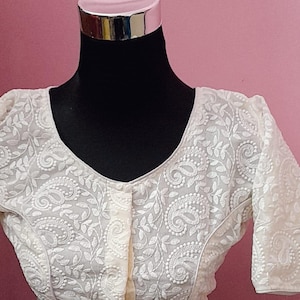 May include: Ivory-colored blouse with intricate floral embroidery. The blouse features a V-neckline, short sleeves, and a button-up front. The embroidery creates a detailed paisley and leaf pattern, adding texture and visual interest to the garment.