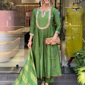 May include: A green, long-sleeved tunic and matching pants set. The tunic features intricate embroidery around the neckline and cuffs, with a pleated design. A striped scarf and a patterned clutch complete the ensemble. The outfit is paired with gold sandals.