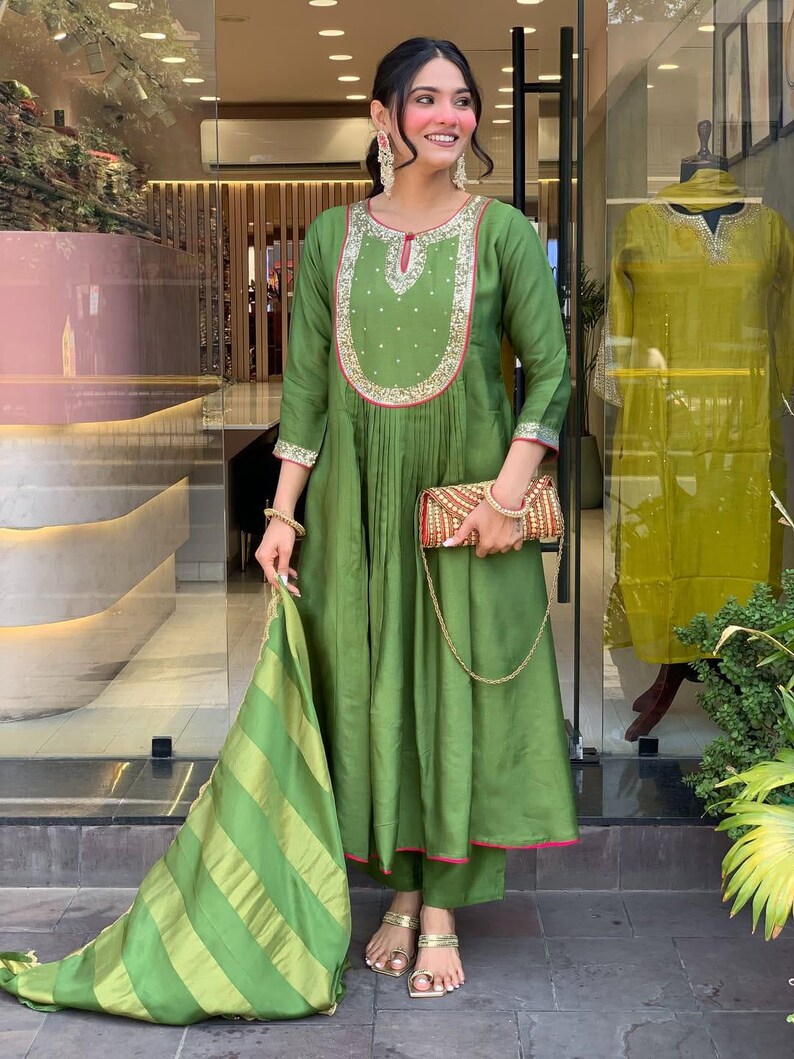 May include: A green, long-sleeved tunic and matching pants set. The tunic features intricate embroidery around the neckline and cuffs, with a pleated design. A striped scarf and a patterned clutch complete the ensemble. The outfit is paired with gold sandals.