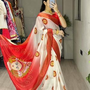 May include: White and red sari with a printed design. The sari features a red border and a large image of a face on the red portion. The white portion has a repeating sun-like design. The sari is draped over a person.