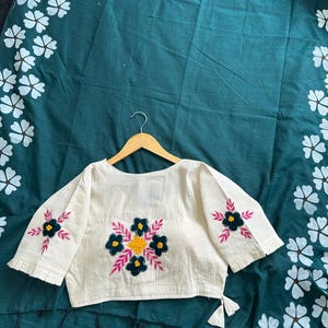 Puede incluir: Top bordado color crema con mangas cortas, con dise&ntilde;os florales en azul marino, amarillo y rosa. La parte superior se muestra en una percha de madera sobre una tela verde oscuro con estampados florales blancos.