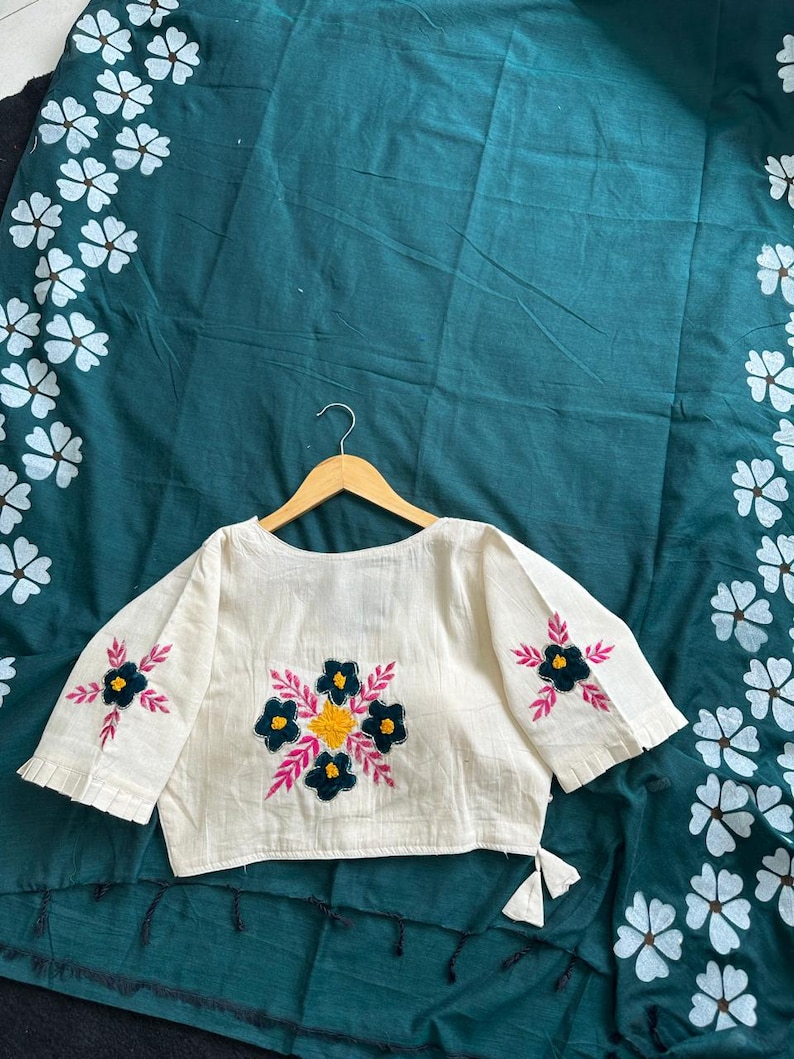 Puede incluir: Top bordado color crema con mangas cortas, con dise&ntilde;os florales en azul marino, amarillo y rosa. La parte superior se muestra en una percha de madera sobre una tela verde oscuro con estampados florales blancos.