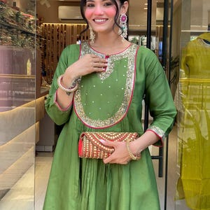 May include: An olive green dress with gold and pink trim, featuring a matching clutch. The dress has a round neckline and long sleeves. The clutch is red with gold embroidery. The woman is wearing large earrings and a bracelet.