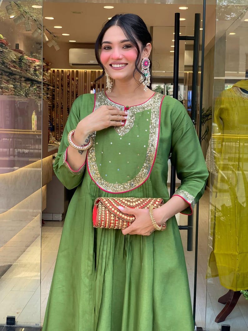 May include: An olive green dress with gold and pink trim, featuring a matching clutch. The dress has a round neckline and long sleeves. The clutch is red with gold embroidery. The woman is wearing large earrings and a bracelet.